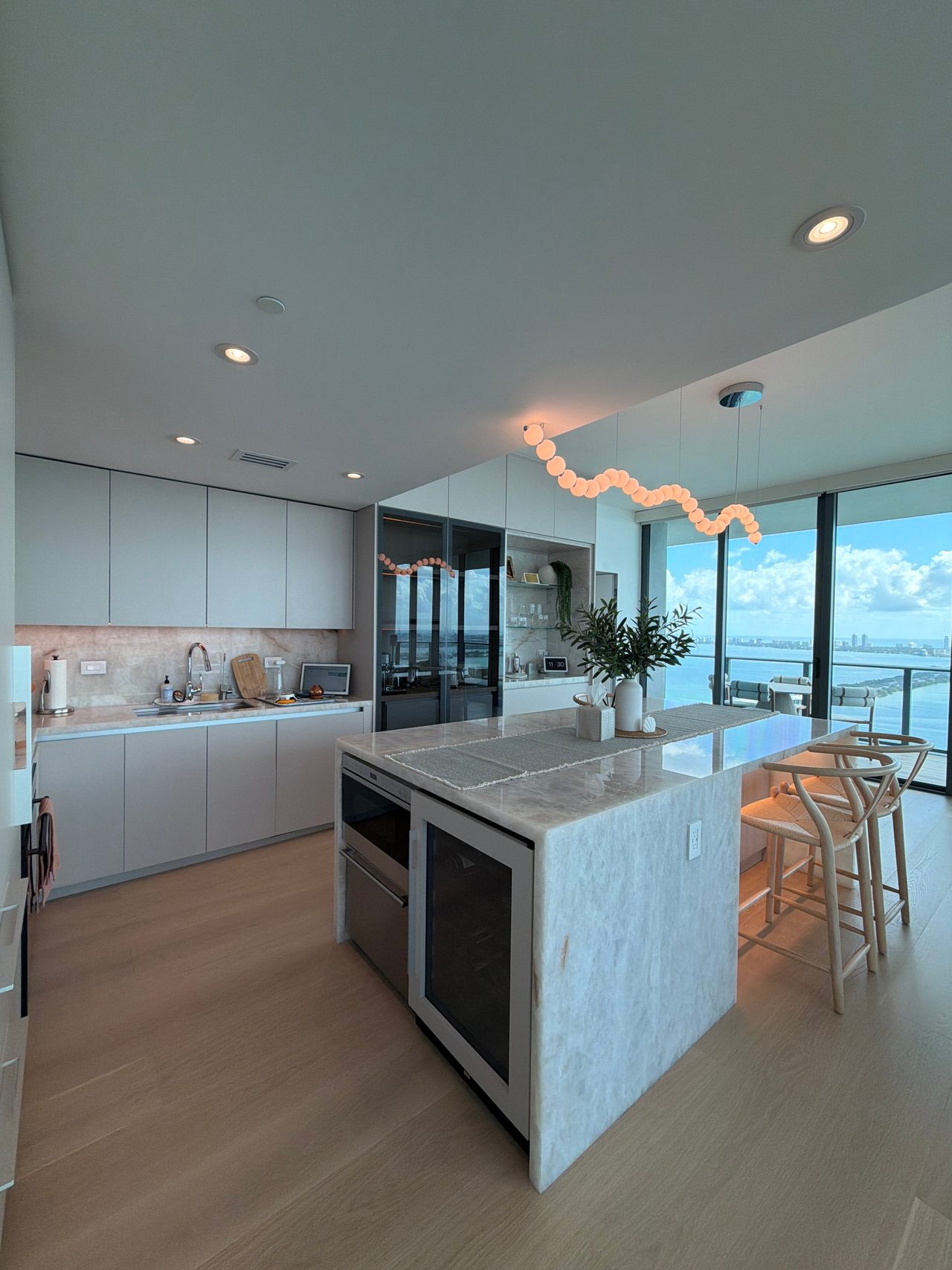 Kitchen with white flat-panel cabinetry, a veined-stone waterfall island holding a wine cooler and oven, woven-seat stools, and a wavy bubble pendant