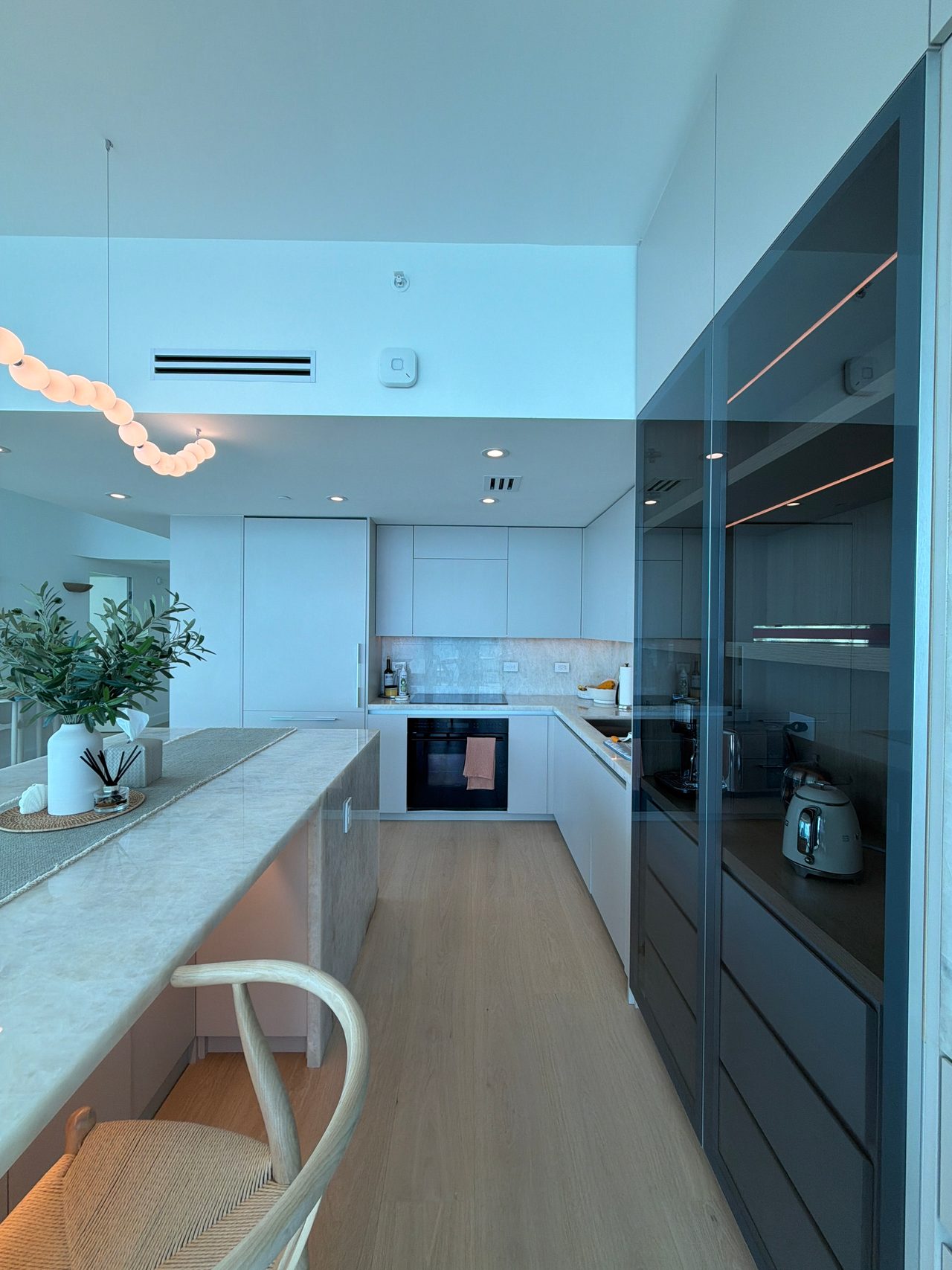 Kitchen with white flat-panel cabinets, a built-in oven, a stone-top island with a woven-seat stool, and a tall smoked-glass pantry cabinet