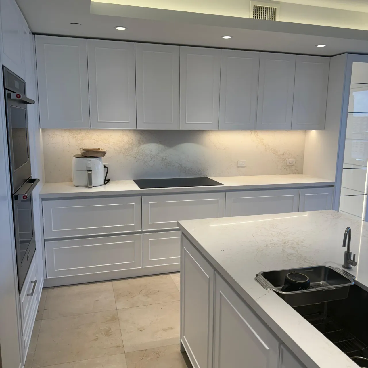 White kitchen with framed-panel cabinets, veined light stone counters and backsplash, a black cooktop, stainless wall ovens and an island sink