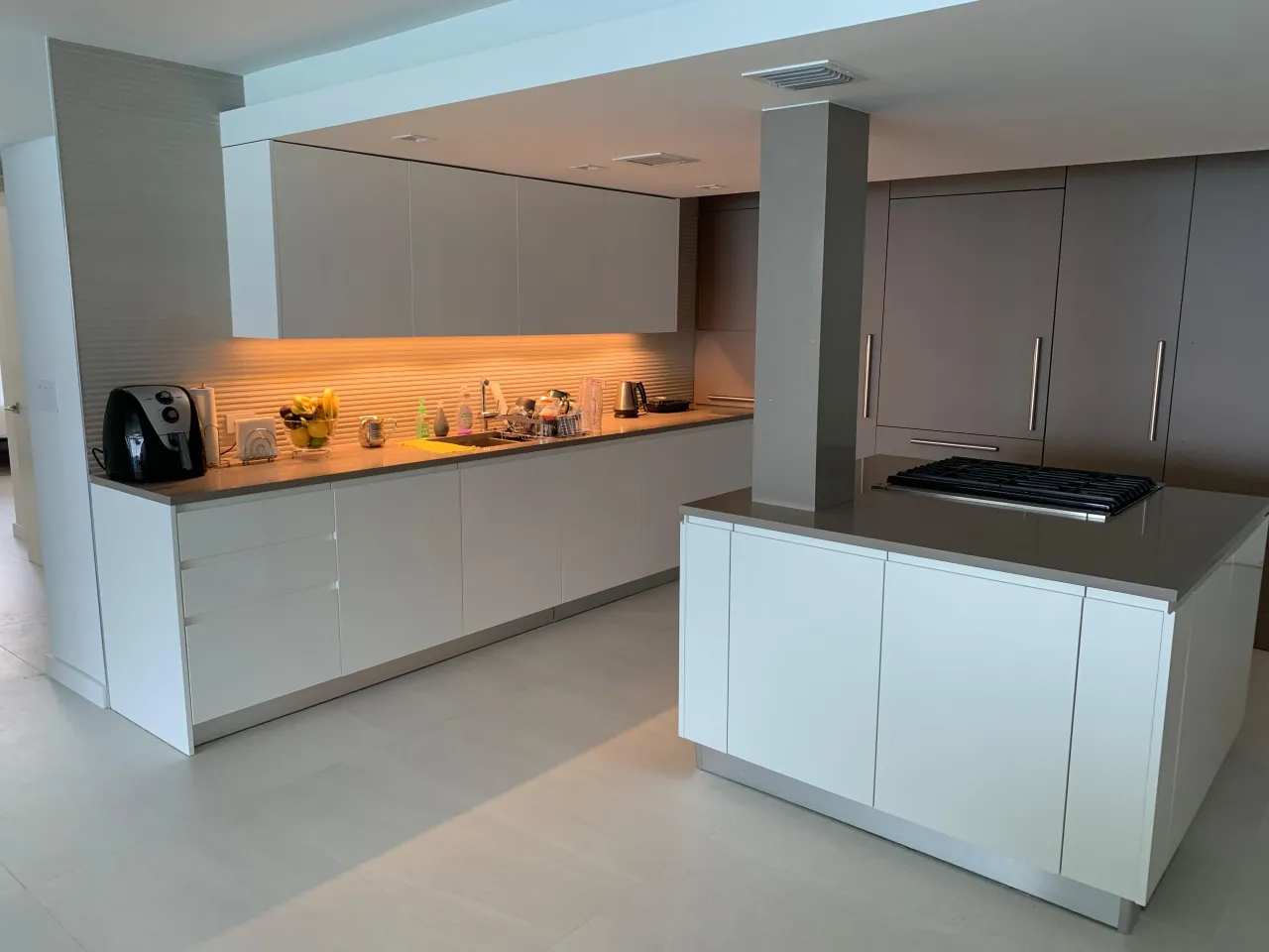 White high-gloss galley kitchen with an L-shaped counter, a separate white island with a black gas cooktop, and taupe cabinetry