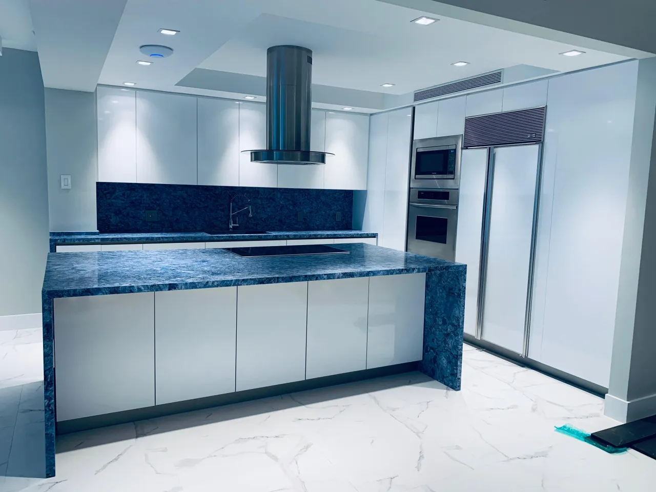 White high-gloss kitchen with a blue-gray stone waterfall island, a cylindrical stainless hood, and a stainless fridge