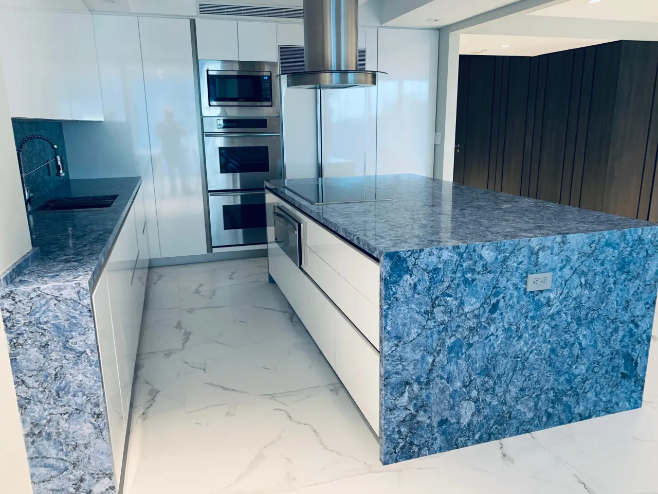 White high-gloss kitchen with a blue stone island counter and backsplash, wall ovens, and stainless appliances