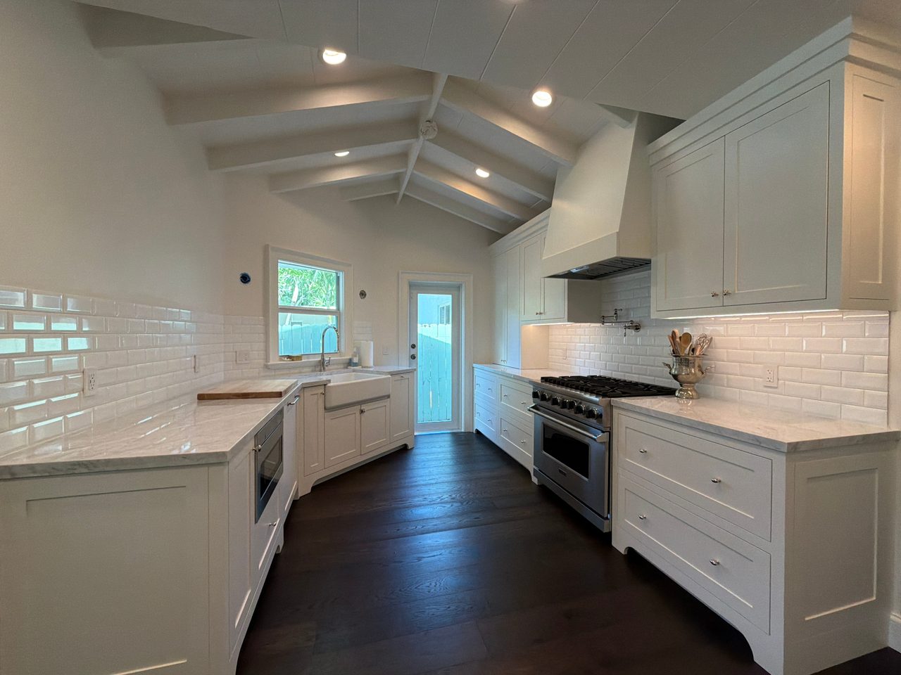 Galley kitchen with white inset shaker cabinets, beveled white tile backsplash, light stone-look counters, and a vaulted beamed ceiling
