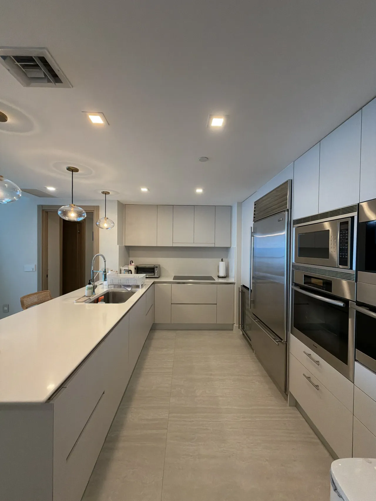 Kitchen with a long white peninsula and sink, light cabinetry, a stainless fridge, and a column of stainless ovens and a microwave