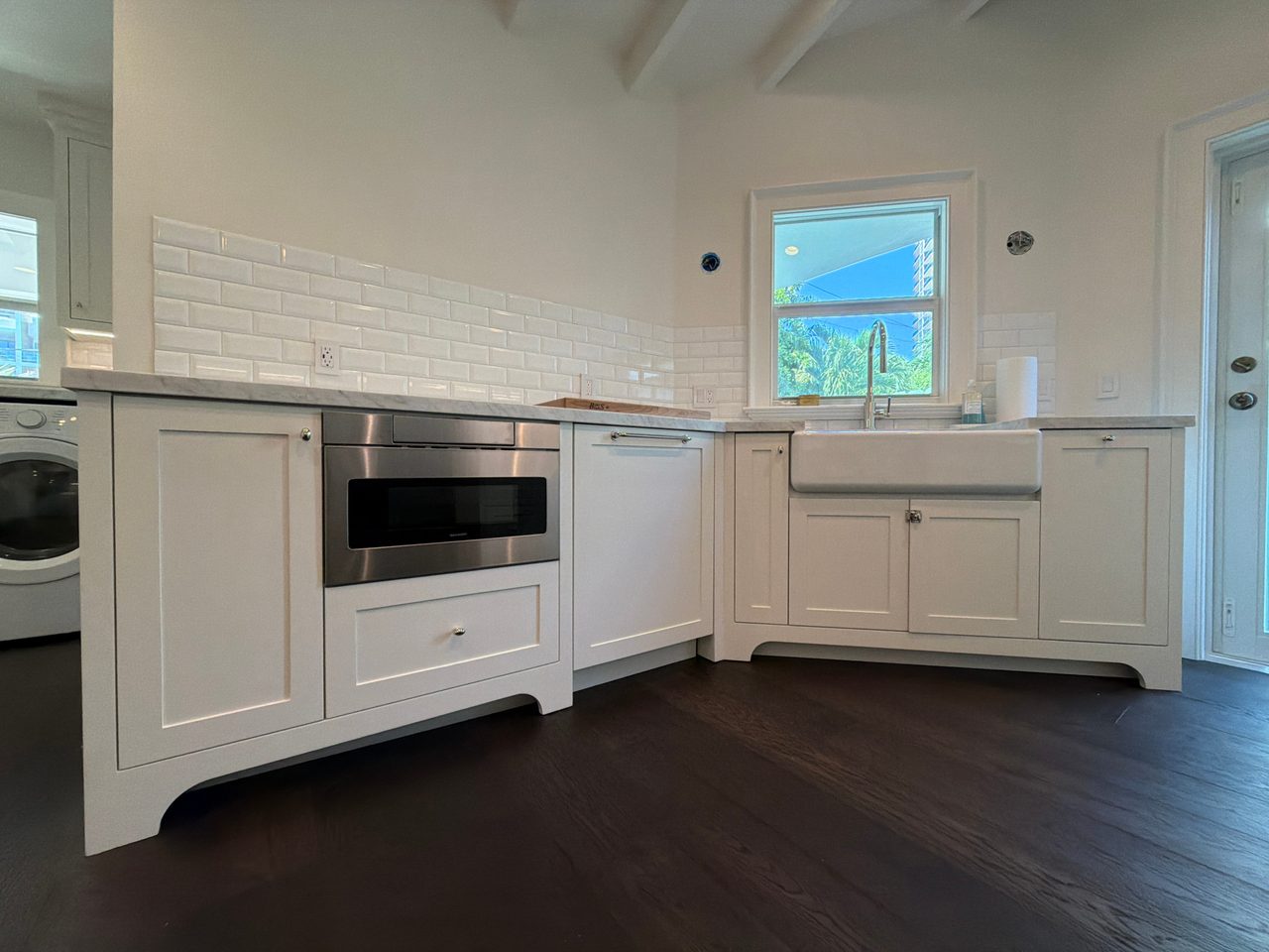 White shaker sink wall with a stainless microwave drawer, apron sink, panel-front dishwasher, and a window above the beveled tile backsplash