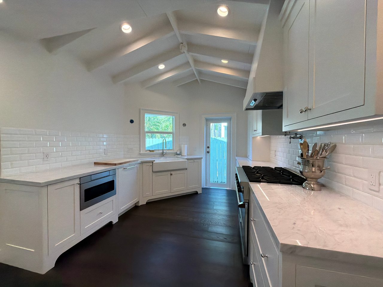 Corner of a white shaker kitchen with a stone-look counter, built-in microwave drawer, apron sink under a window, and a hood above the range