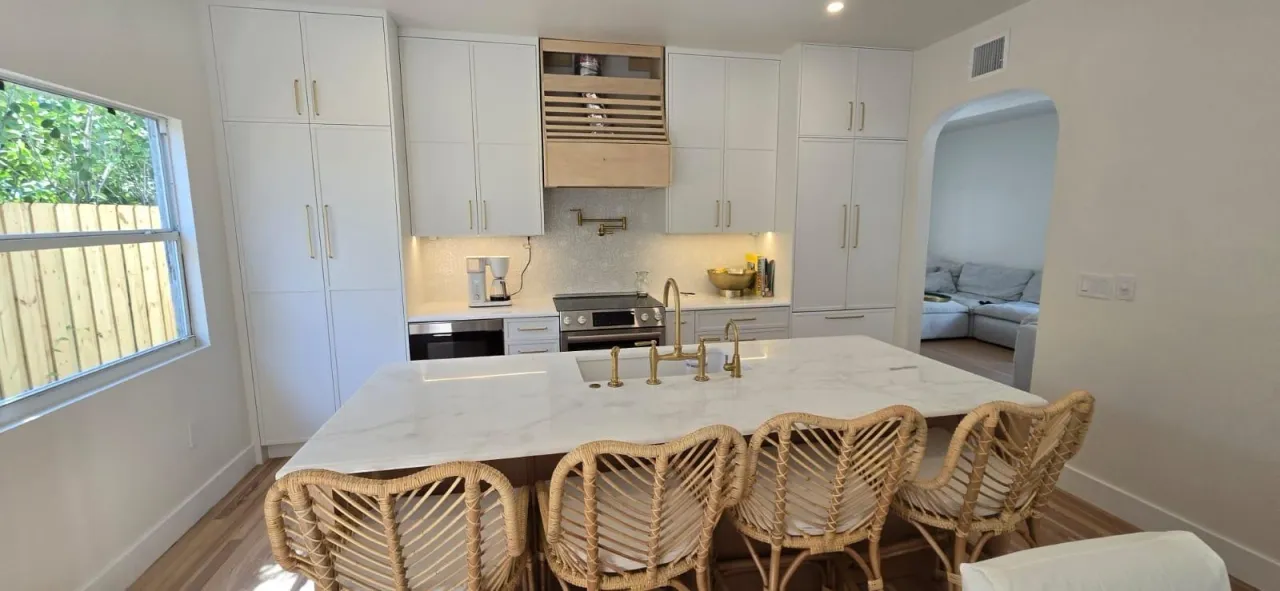 White shaker kitchen with rattan bar stools, a stone island, gold-tone fixtures, and a wood plank range hood cover above the cooktop