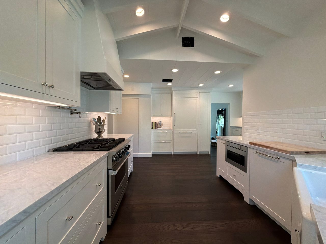 Long white shaker kitchen with stone-look counters, a gas range, under-cabinet lighting, and a paneled refrigerator at the far end on dark wood floors