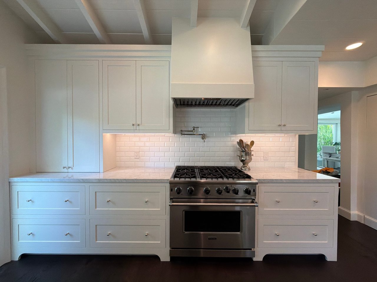 White shaker cabinetry around a stainless range with a painted hood, pot filler, and beveled white tile backsplash under exposed ceiling beams