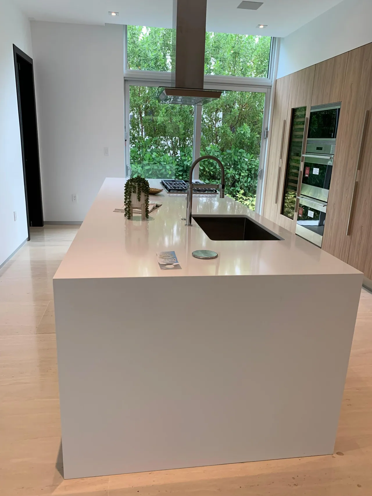 White stone waterfall island facing floor-to-ceiling windows with a garden view, with light wood tall cabinets alongside