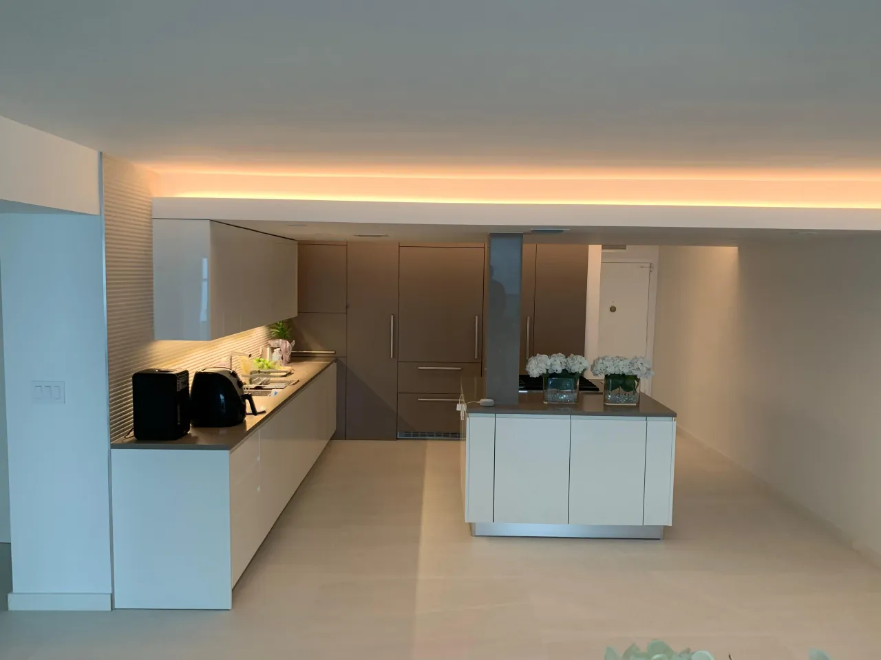 White and taupe kitchen with a glowing ceiling cove light strip, a white island with a gray counter, and taupe tall cabinets with vertical bar pulls