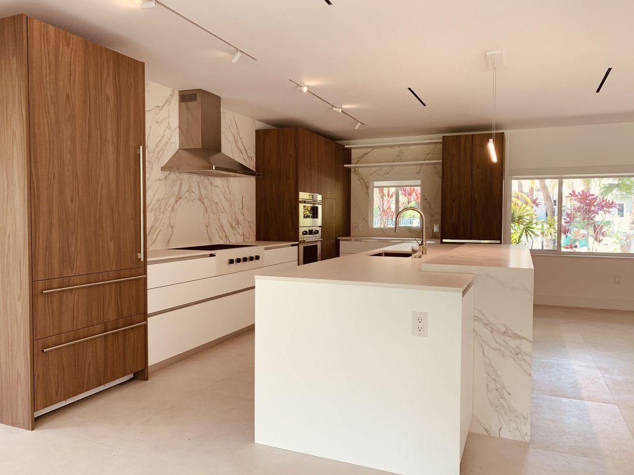Kitchen with a white waterfall island, a dark wood cabinet wall, a veined-stone slab backsplash, and a window