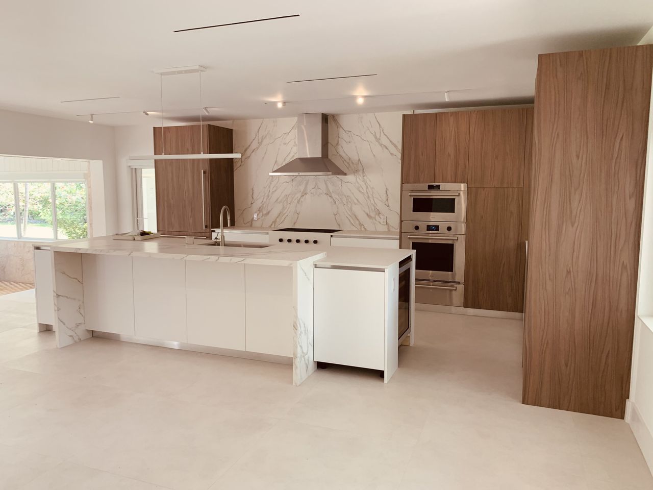 White waterfall island kitchen with a full veined-stone slab backsplash, a stainless range hood over the cooktop, and dark wood cabinetry on the side wall