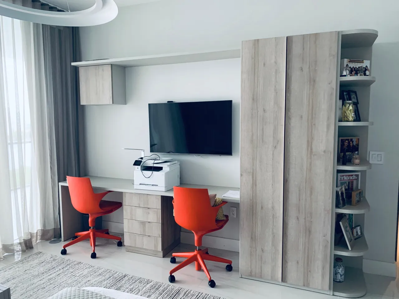Bedroom desk run in wood-grain with a central drawer bank under a wall-mounted TV, an upper cabinet, orange task chairs and a wardrobe with corner shelves