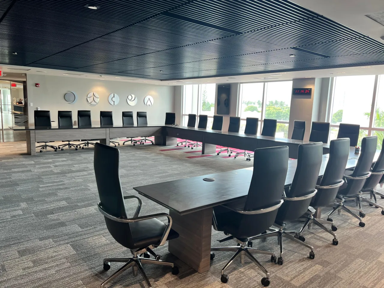 Large boardroom with a wraparound conference table on wood-grain panel bases, black high-back chairs, a slatted ceiling and floor-to-ceiling glazing