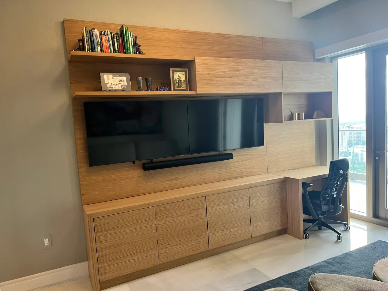 Built-in wood-grain media wall with a mounted TV and soundbar, upper cabinets, open display shelves and a return desk with a black task chair