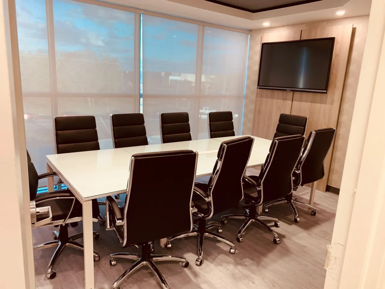Small conference room with black leather-look chairs around a white table, wall-mounted TV on a light wood panel accent wall