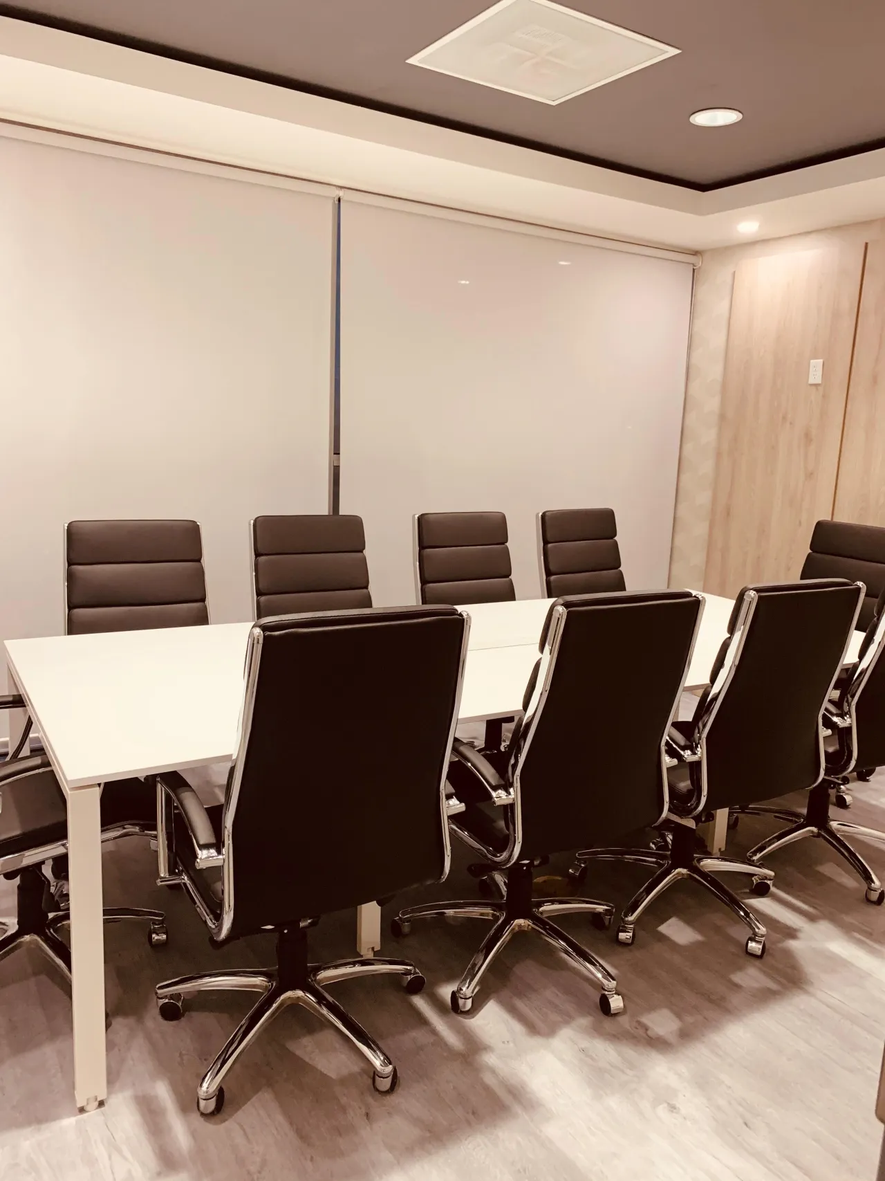 Conference room with a white table, black leather-look chairs on chrome bases, a wood-grain panel wall and a roller-shaded window under a tray ceiling