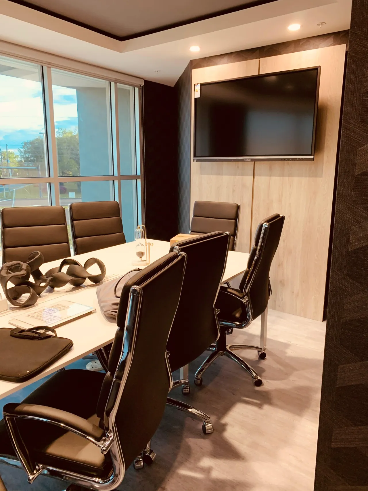 Small conference room with a white table, black leather-look chairs, and a wall-mounted TV on a light wood accent panel, decorative objects on the table