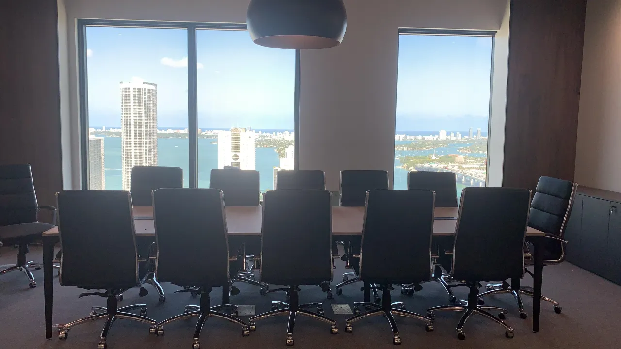 Conference room with a wood table, black leather-look chairs, a large black pendant light, and floor-to-ceiling windows overlooking a bay and city skyline