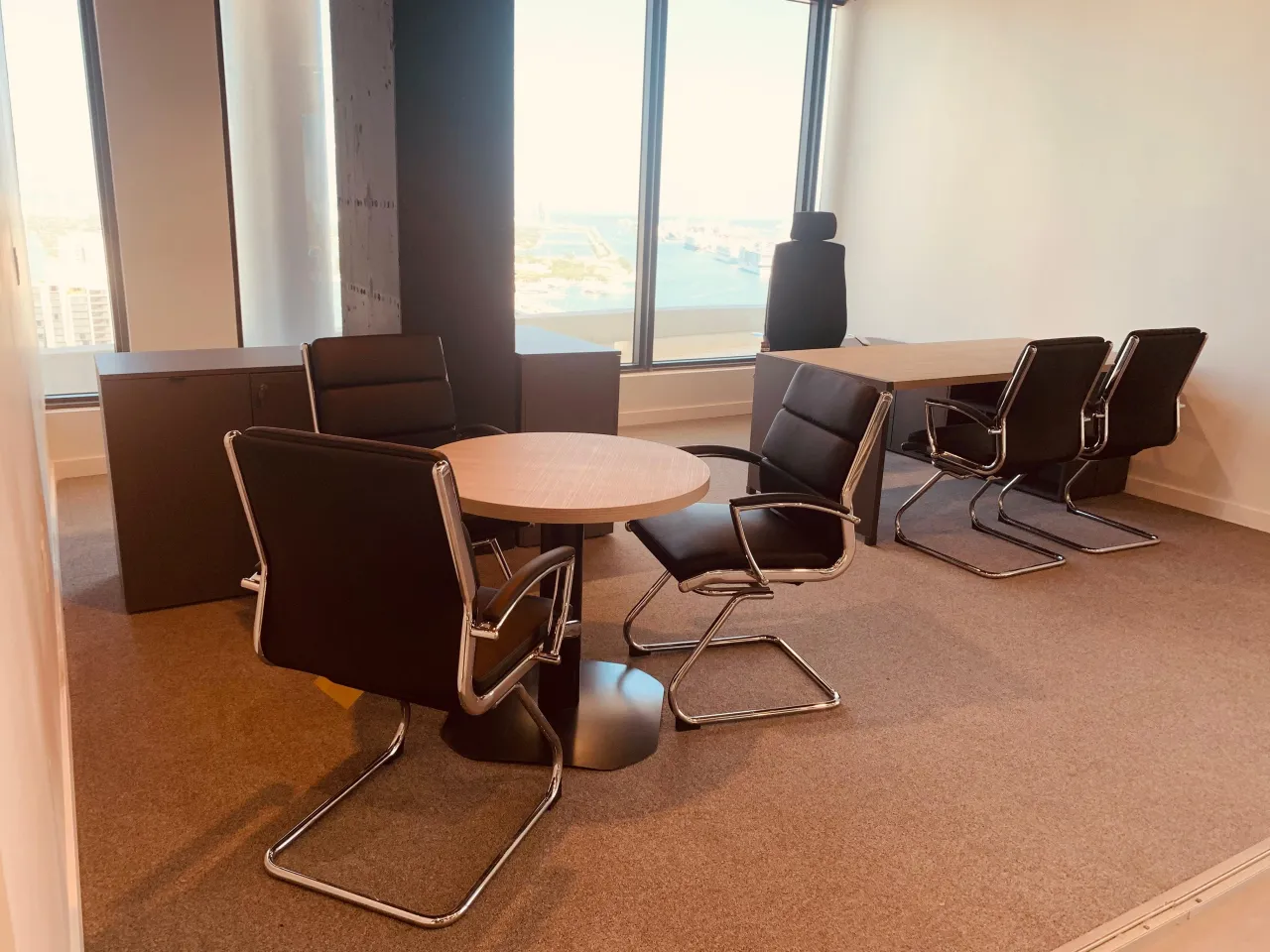 Corner office with a round wood-grain meeting table, black leather-look cantilever chairs, a wood-grain desk and dark storage credenzas on carpet tile