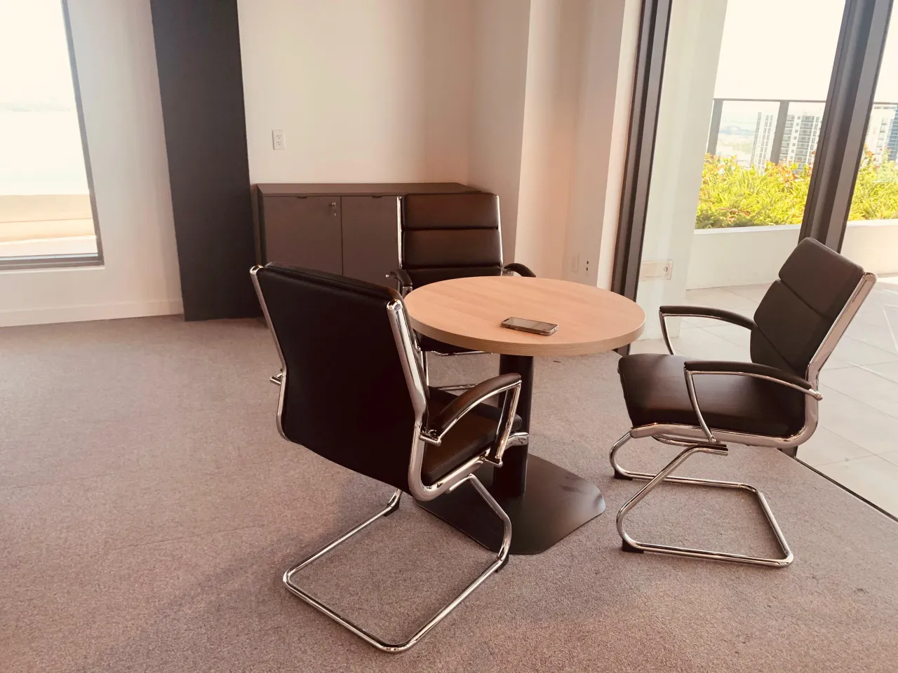 Meeting corner with a round wood-grain table on a black pedestal base, black leather-look cantilever chairs and a dark low storage cabinet by the glazing