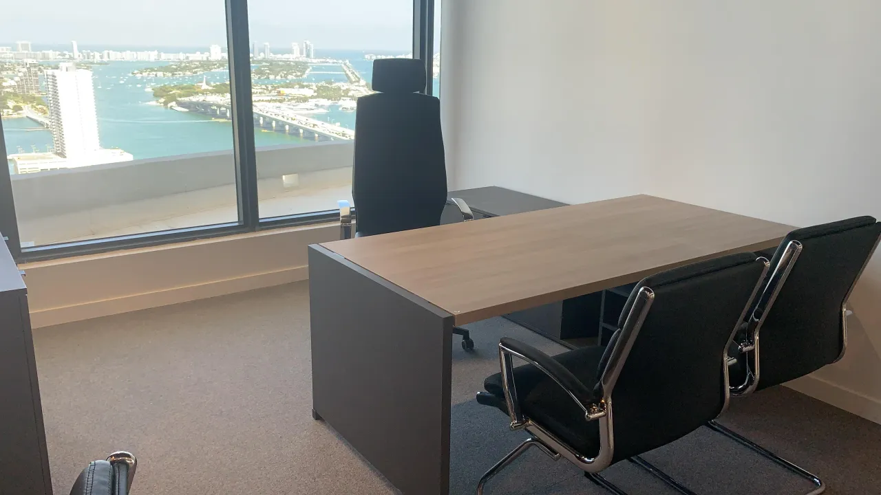 Corner office with a wood desk, black leather-look guest chairs, and floor-to-ceiling windows showing a panoramic waterfront city view