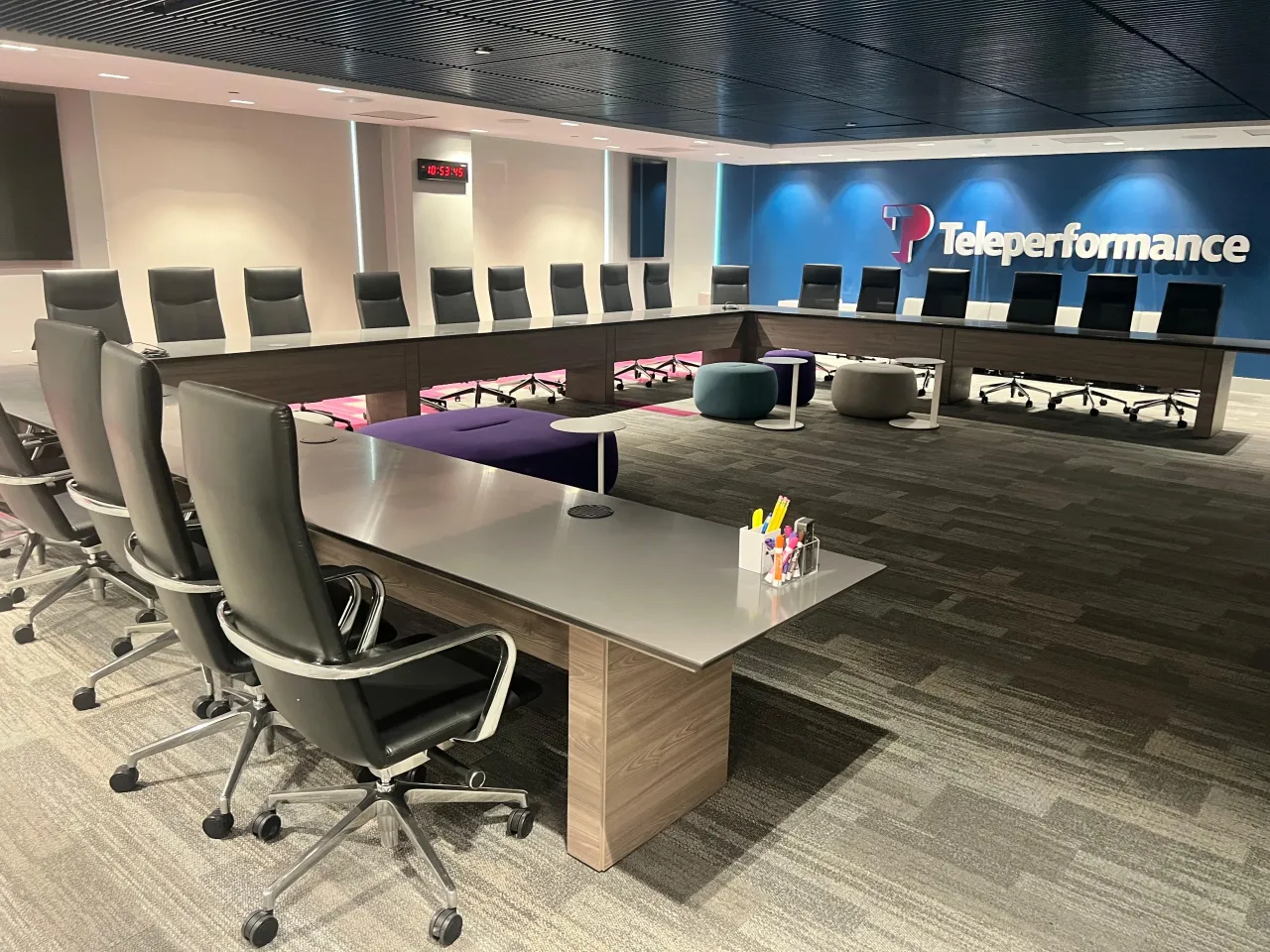 Corporate boardroom with a U-shaped conference table on wood-grain panel bases, rows of black high-back chairs, ottoman seating and a slatted ceiling