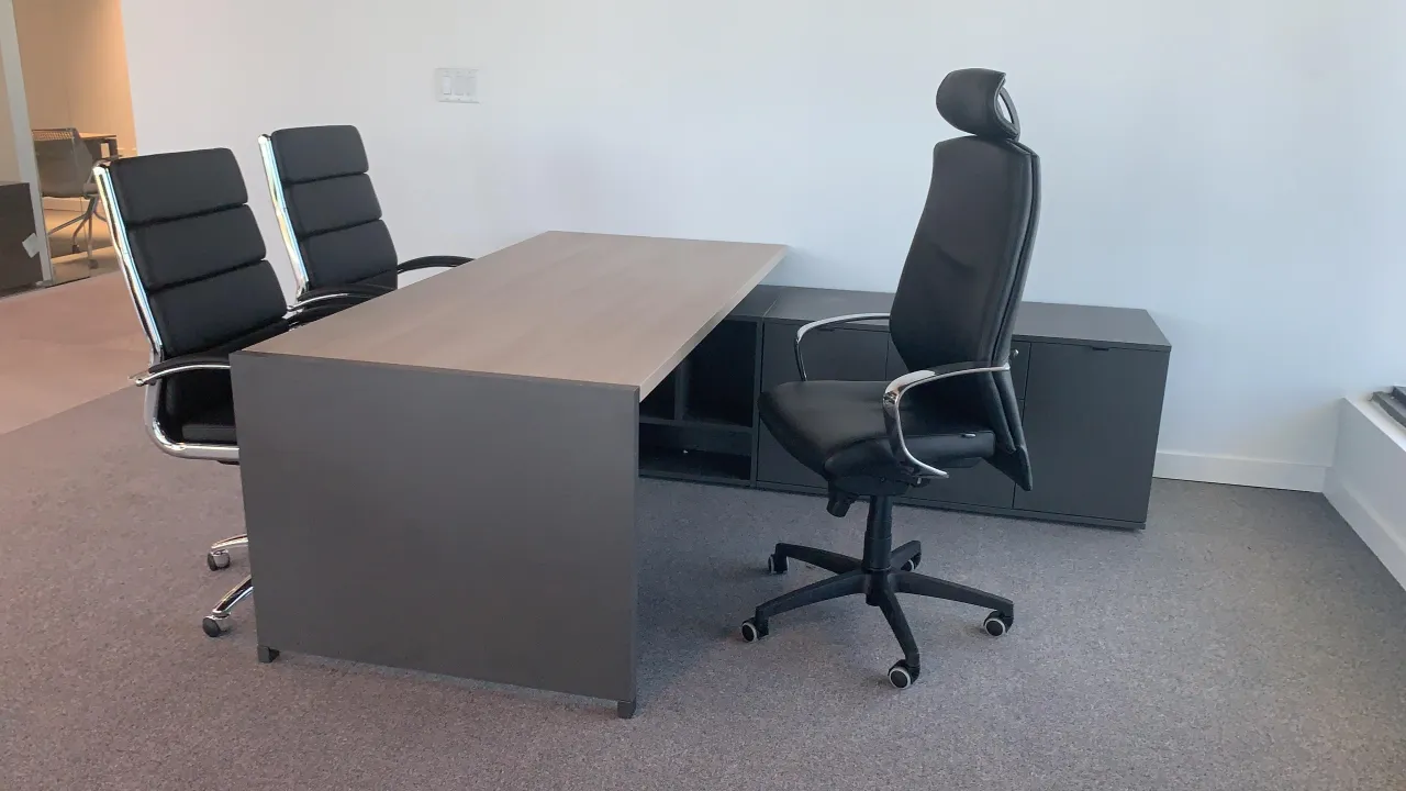 Executive office with a wood-grain desk on a dark panel base, a dark low credenza behind it, a high-back task chair with headrest and two guest chairs