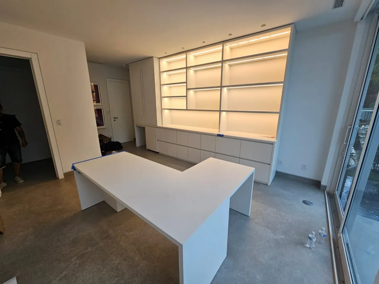 Home office under installation with a white built-in wall unit, LED-lit open shelving over a long drawer bank, and a freestanding white L-shaped desk