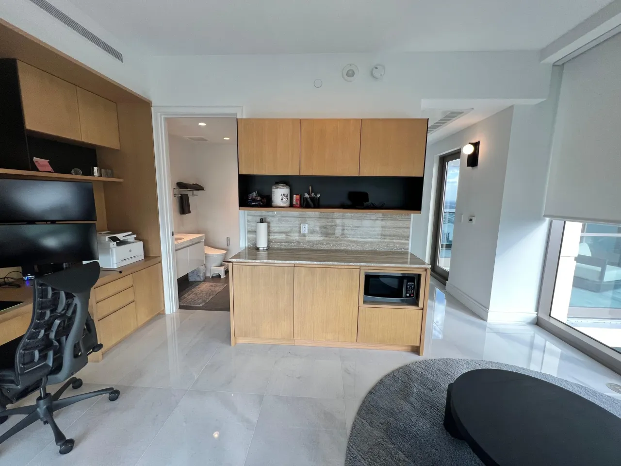 Home office kitchenette with wood-grain cabinets, a veined-stone counter and backsplash, a dark shelf niche, built-in microwave and a bath doorway beyond