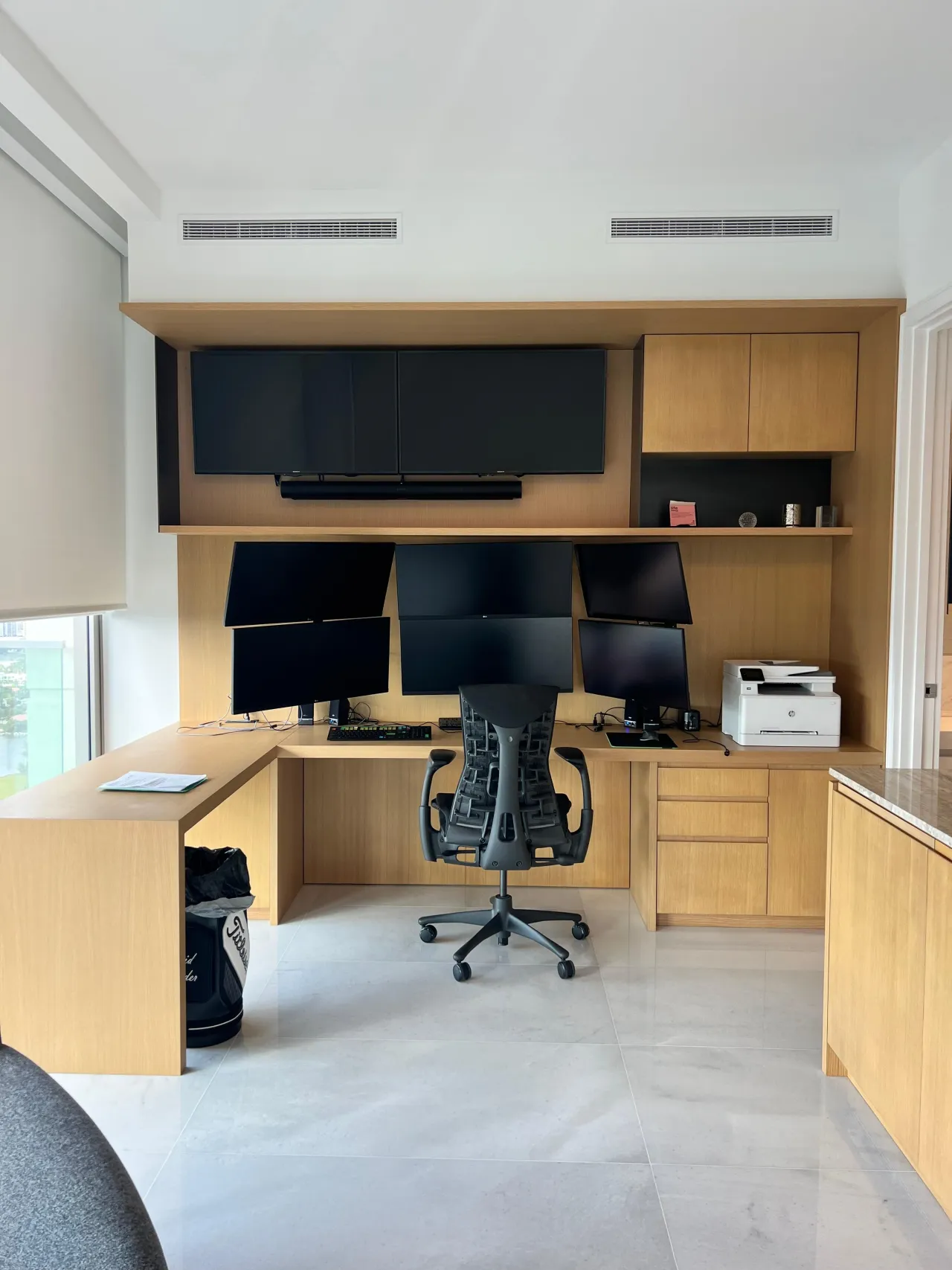 Home office with a light wood built-in U-shaped desk, upper flat-panel cabinets with a soundbar, and a multi-monitor setup with a black task chair