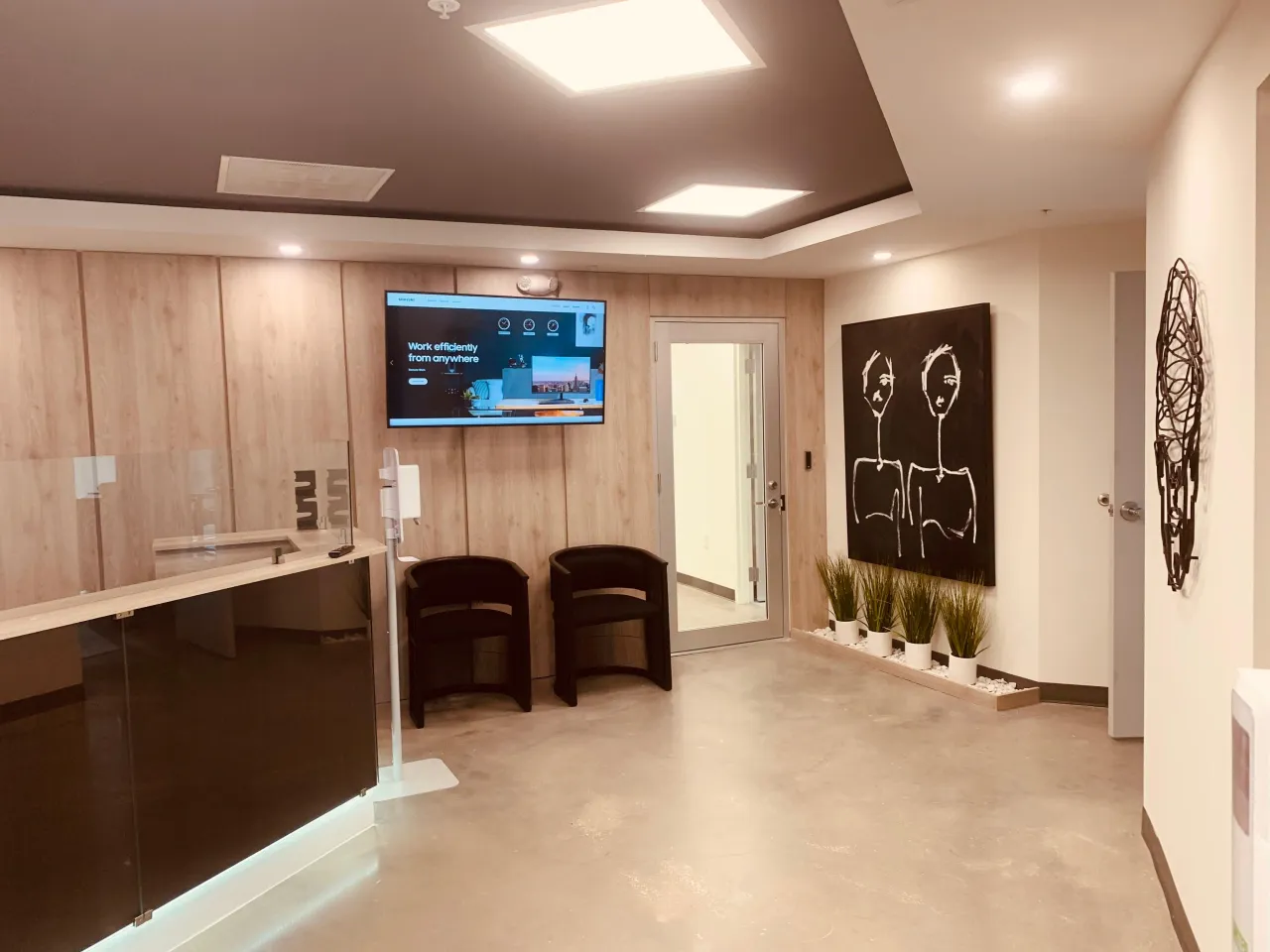 Lobby with a wood-grain panel wall, a wall-mounted display screen, a dark reception counter with a glass screen, black barrel chairs and a planter niche