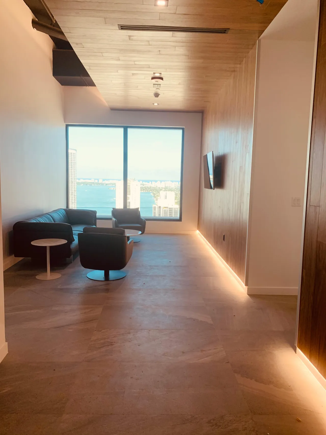 Lounge corridor with a wood-plank ceiling and wood-clad wall with concealed LED, a wall-mounted screen, dark sofa and swivel chairs by a corner window