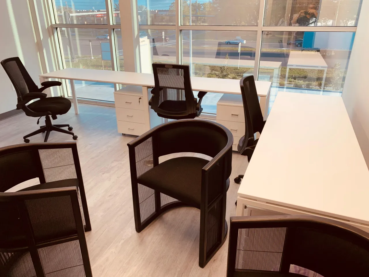 Open office area with white desks in a row along floor-to-ceiling windows, black task chairs and black barrel guest chairs