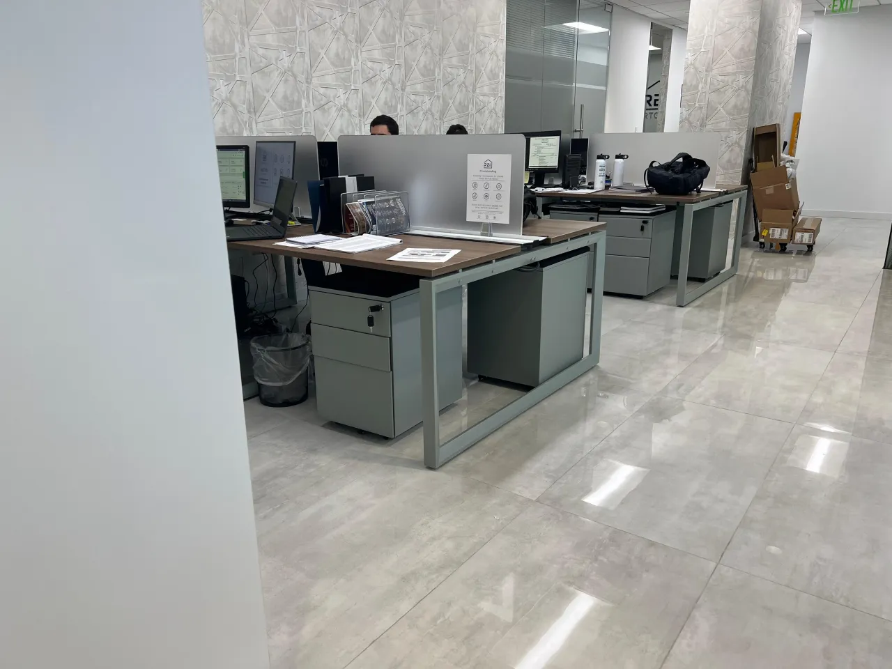 Open office workstation area with wood-top desks on gray frames, pedestal file cabinets, monitors, patterned wall visible in background corridor