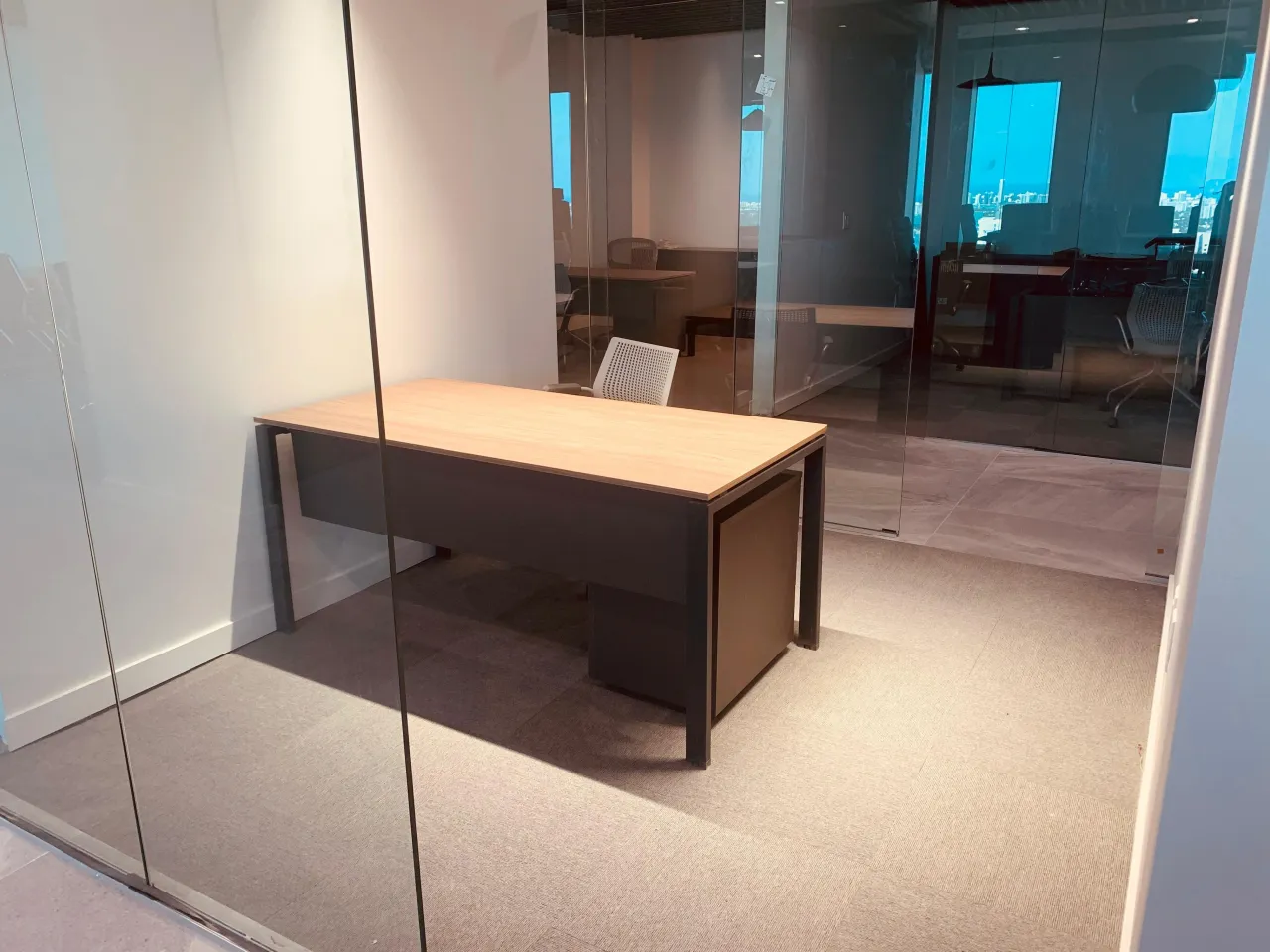 Private office behind a full-height glass partition with a wood-grain desk on a dark metal frame and a matching pedestal cabinet, on carpet tile