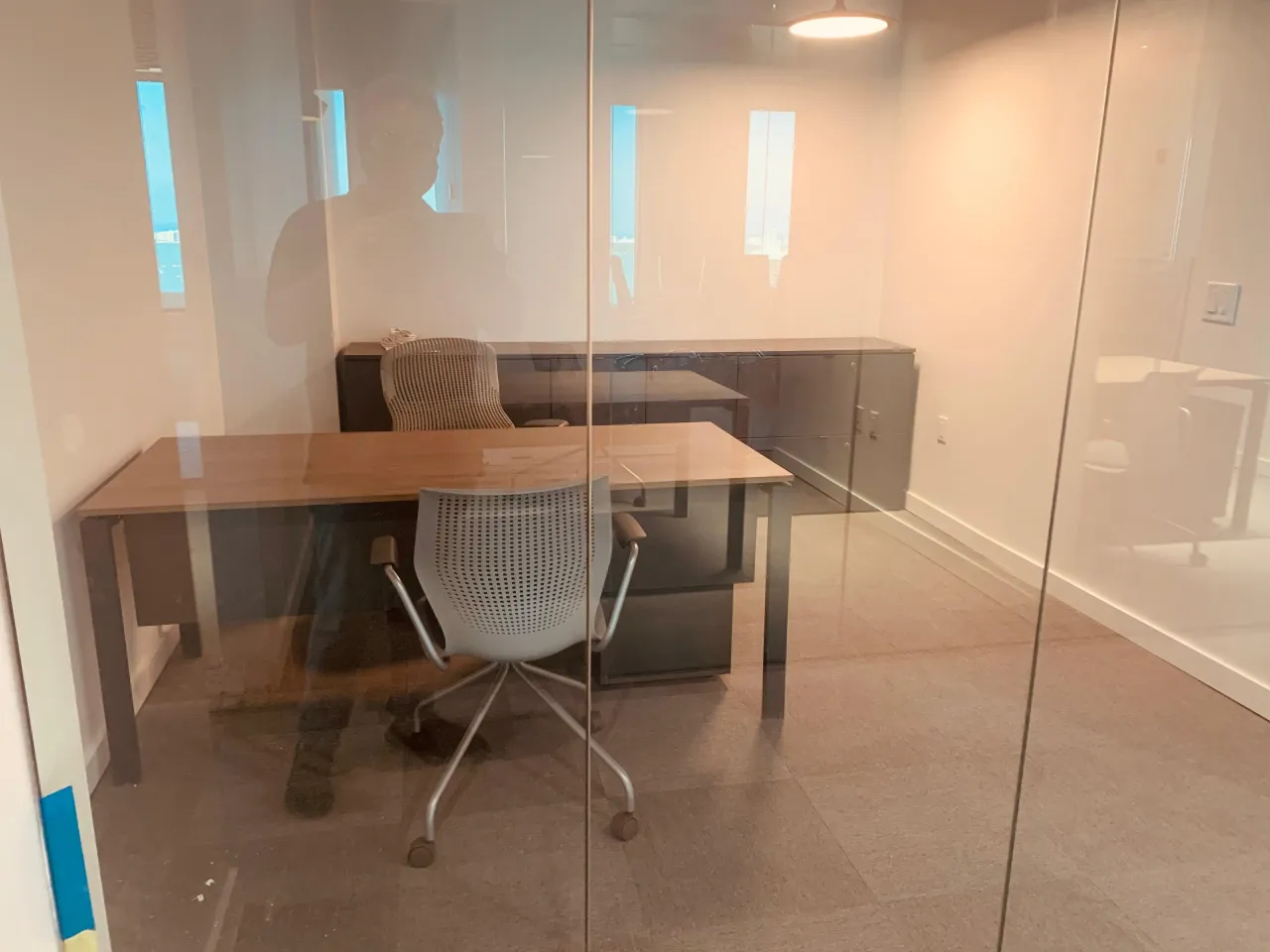 Private office behind glass partition with a wood desk, task chairs, storage cabinets along the wall, seen through reflective glass