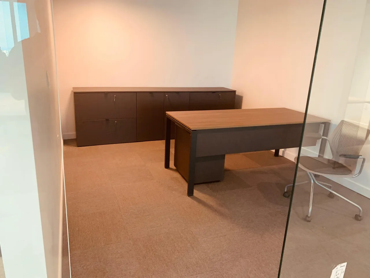 Private office behind a glass partition with a wood-grain desk on a dark metal frame and a dark lateral credenza run along the back wall, on carpet tile