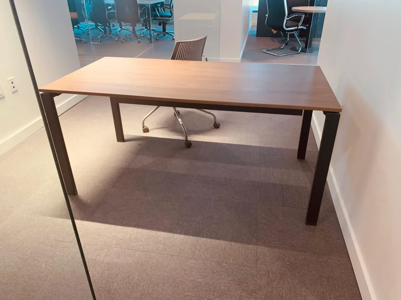 Small private office with a light wood desk on dark metal legs and a single task chair, glass partition wall to the corridor