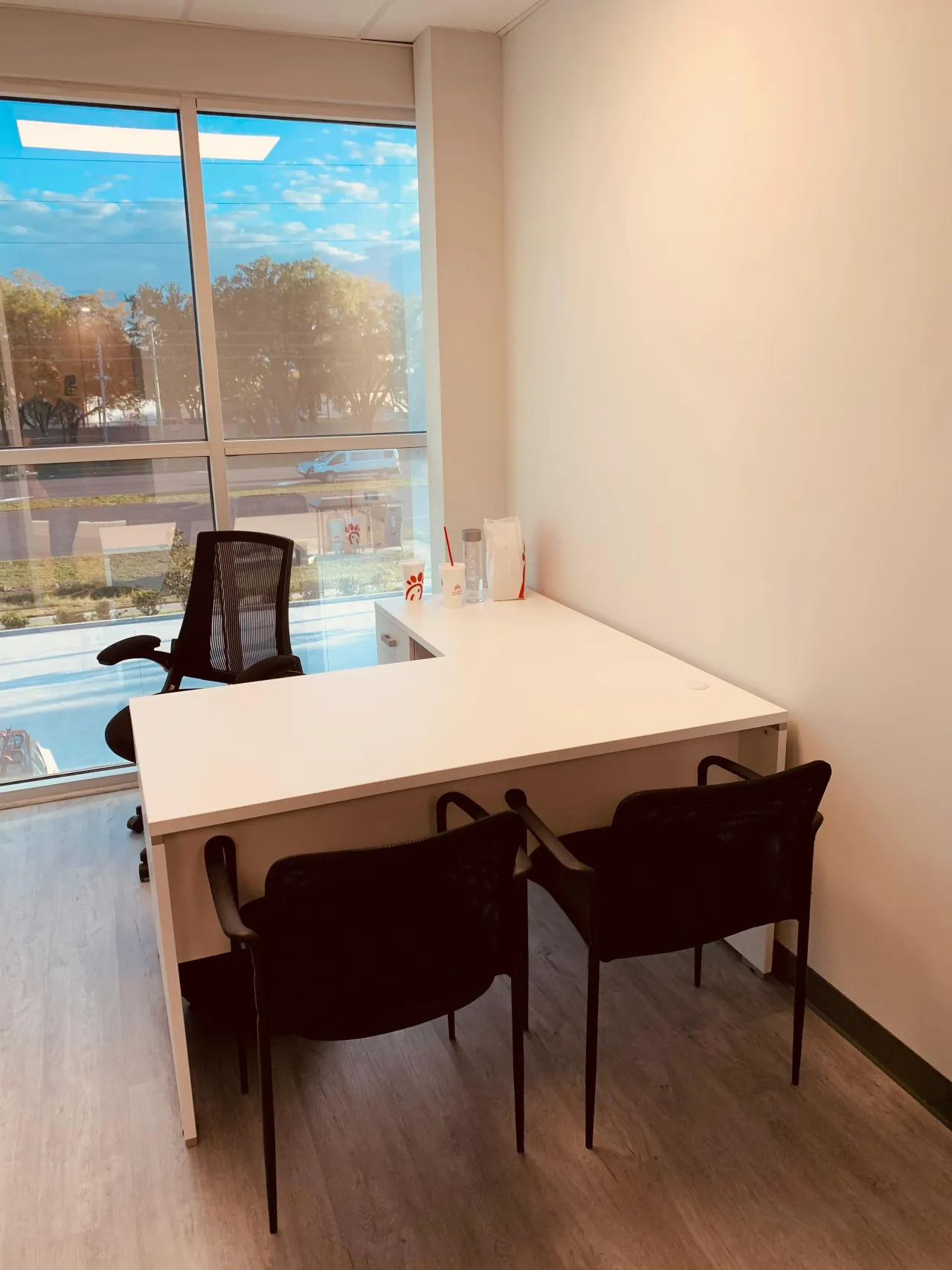 Private office with a white L-shaped desk beside a full-height window, a black mesh task chair, two black guest chairs and drink cups on the return