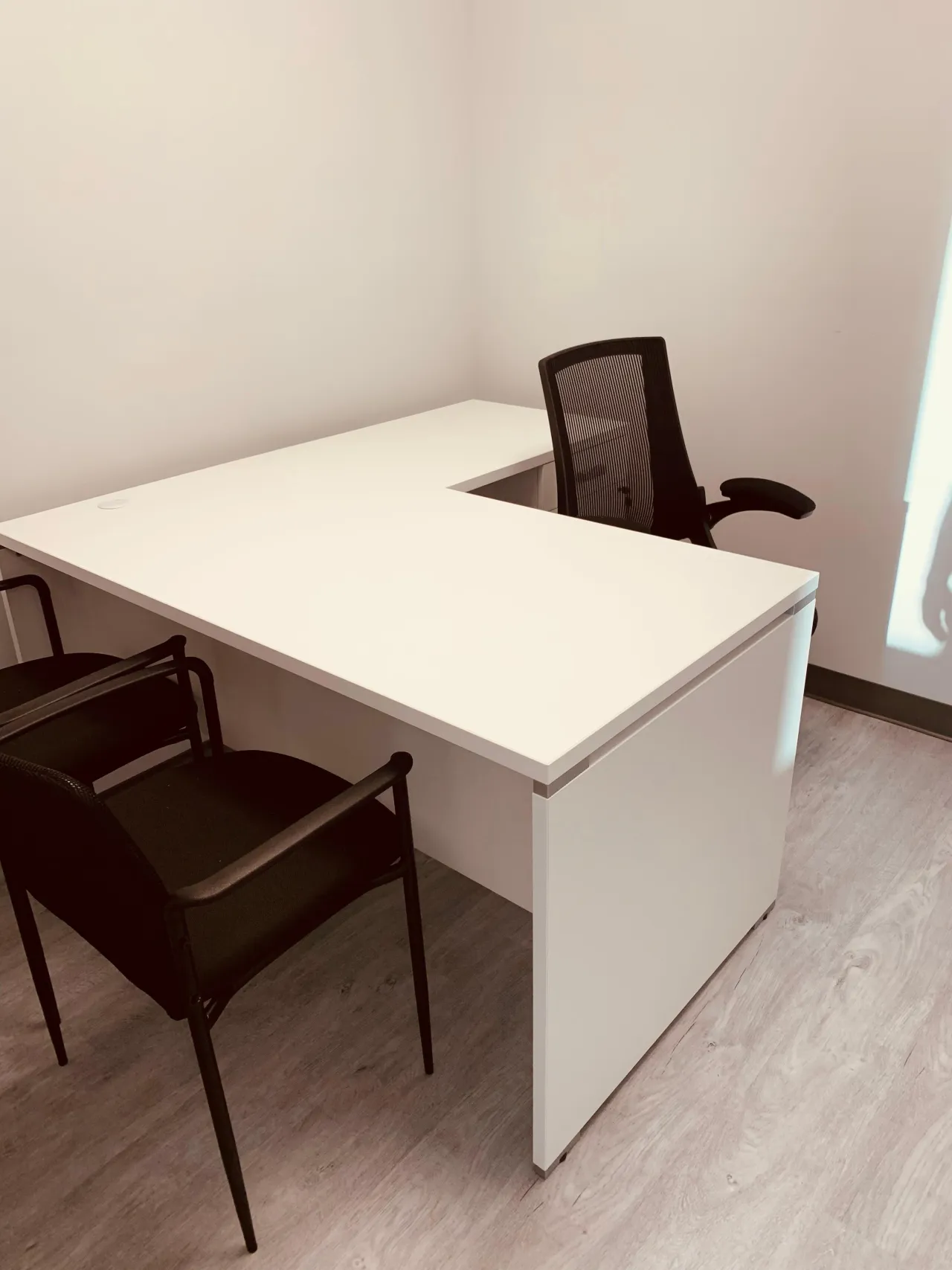 Small private office with a white L-shaped desk over a pedestal cabinet, a black mesh task chair and two black guest chairs on vinyl plank flooring