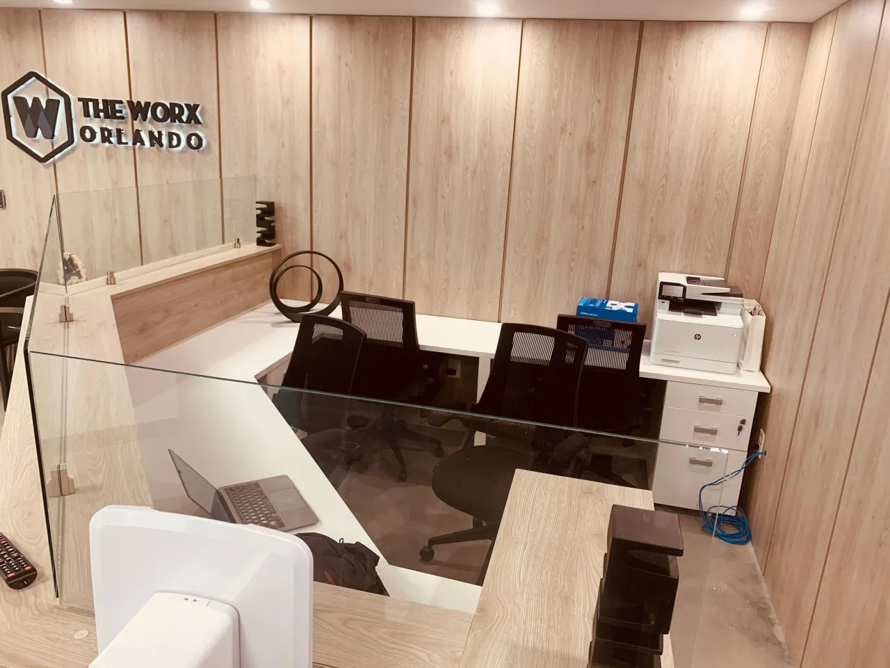 Reception area with light wood-panel walls, a glass partition desk, black task chairs and a printer station