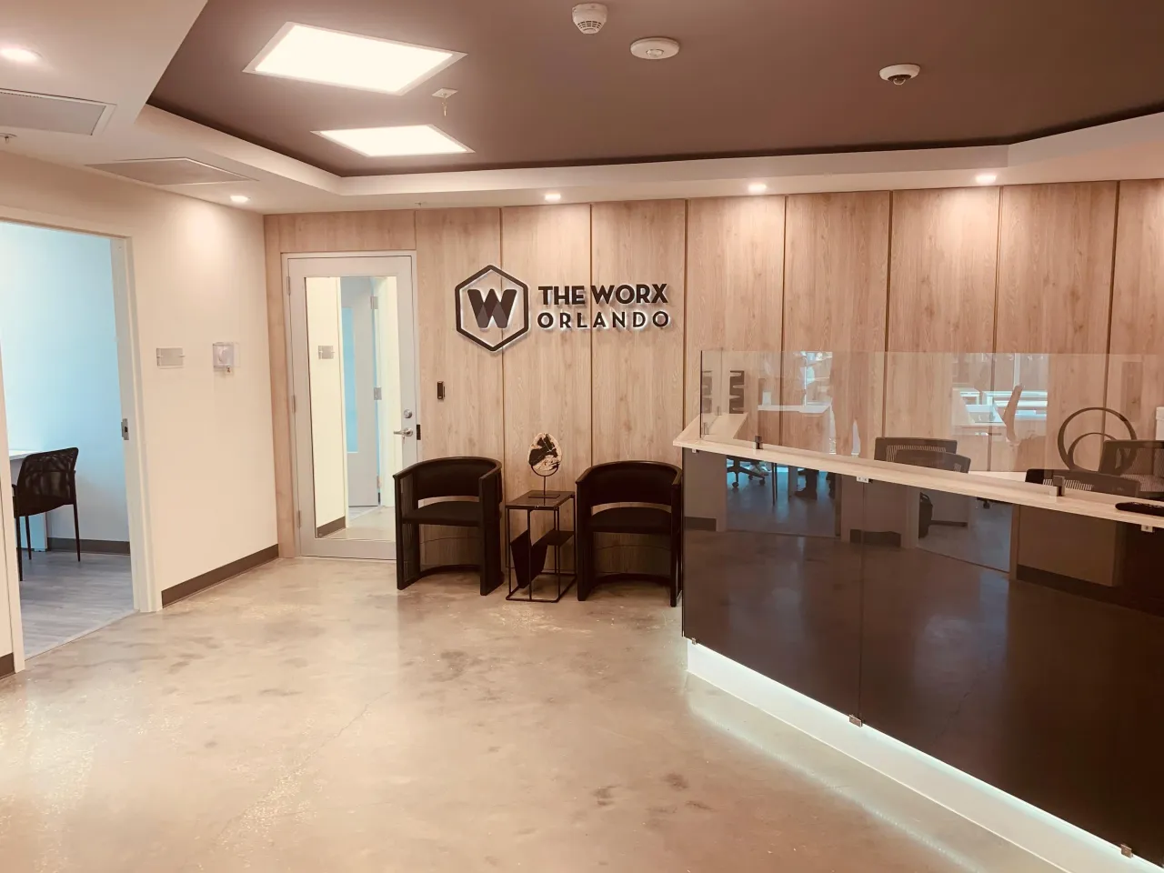Reception area with a vertical wood-grain panel wall, a dimensional logo sign, a dark reception counter with glass screens and black barrel guest chairs