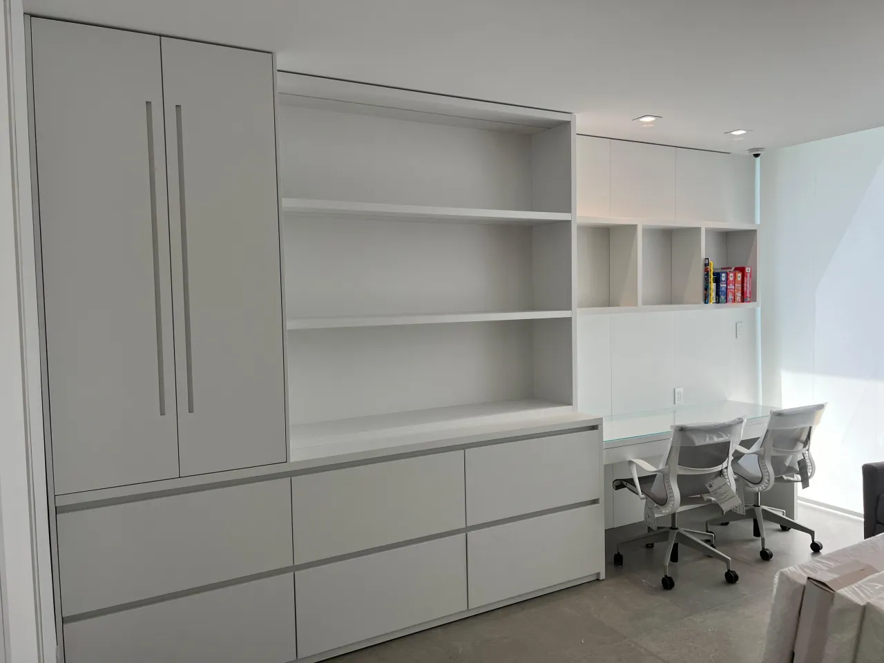 White built-in wall unit with a tall cabinet, open shelving and a long drawer bank, beside a glass-topped desk return with two white mesh task chairs
