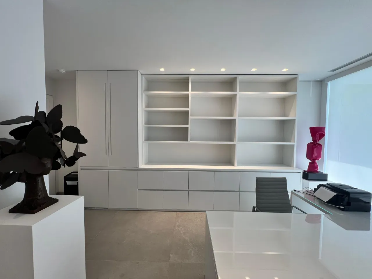 White built-in wall unit with a tall cabinet, an empty open shelving grid and a long drawer bank, beside a white desk with a glass top and a task chair