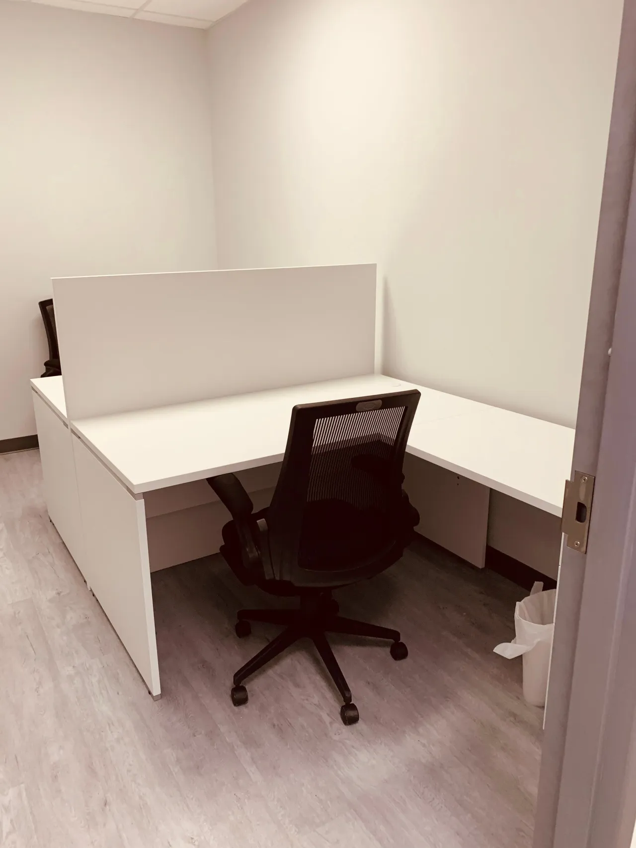 Small office with a white desk and black mesh chair divided by a white privacy panel from a second workstation with matching desk and chair
