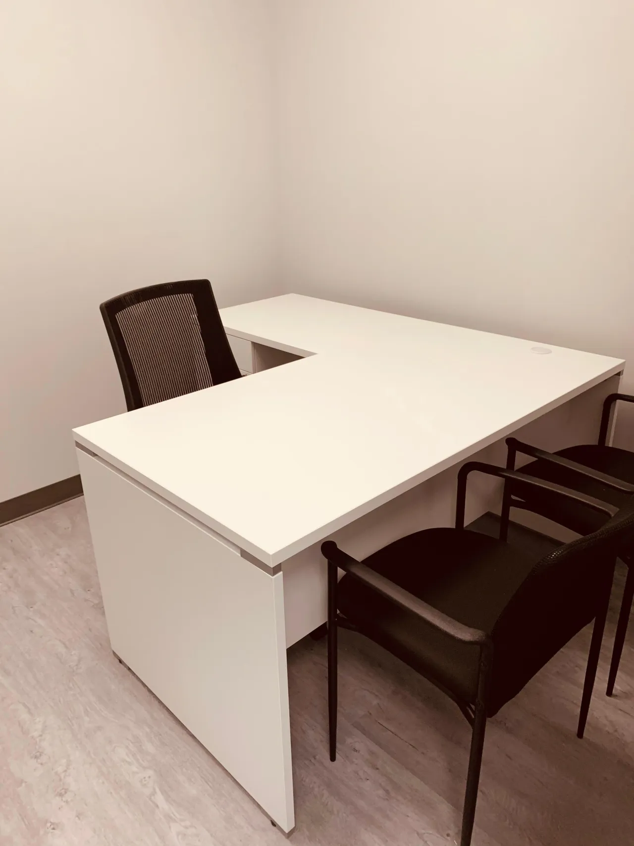 White L-shaped desk with a pedestal drawer unit, a black mesh task chair behind it and two black guest chairs opposite, on vinyl plank flooring