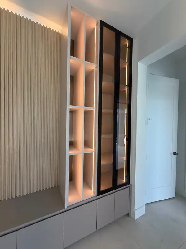 Backlit white grid shelving column next to a tall black-framed glass cabinet, over low gray cabinet fronts, wood-grain slat wall at left