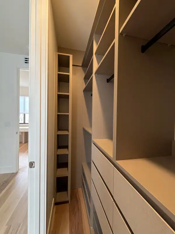 Walk-in closet in beige with a tall narrow cubby tower, open shelves, black hanging rods and a bank of flat-front drawers on a wood-grain floor